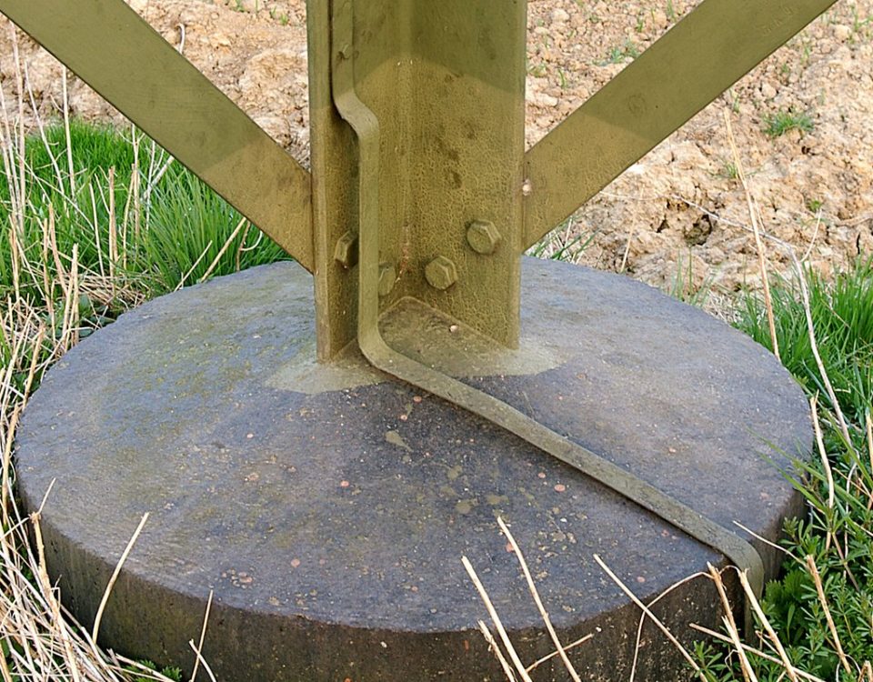 Galvanized steel transmission tower leg bolted to a concrete foundation, with a flat earthing strip running down the leg into the ground